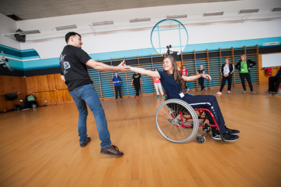 Une jeune fille dans un fauteuil roulant danse avec un homme debout. Ils se tiennet par la main.