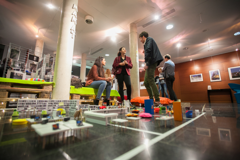 Trois étudiants en train de discuter vus en contre-plongée. À leurs pieds, des meubles miniatures évoquant une bibliothèque.