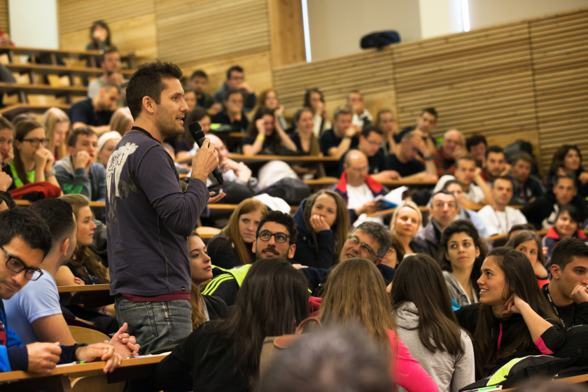 Un amphithéâtre plein dans lequel un étudiant grec debout tient un micro.