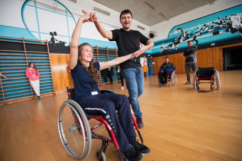 Une jeune fille dans un fauteuil roulant danse avec un homme debout. Ils se tiennet par la main.