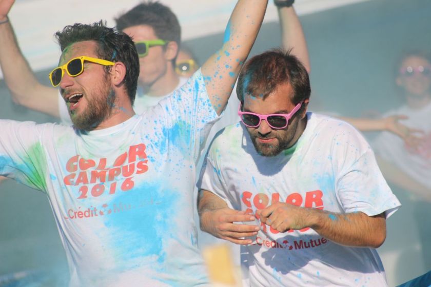 Deux étudiants avec des tee shirts blancs constellés de poudre colorée et des lunettes de couleur font des gestes enthousiastes.