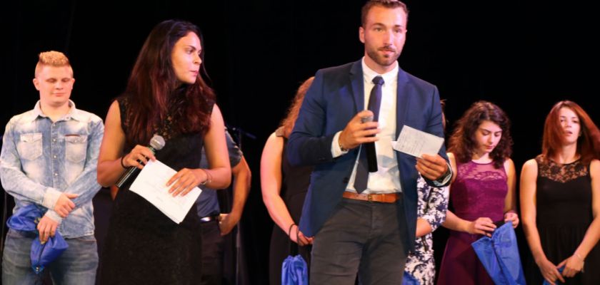 Antoine Mairot tenant un micro sur une scène, derrière lui des étudiants en tenue de gala qui tiennent des sacs cadeaux dans les mains.