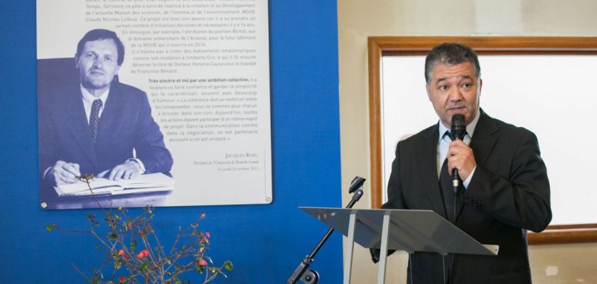 Jacques Bahi devant un pupitre tenant un micro. En arrière plan, un portrait de Claude Oytana.
