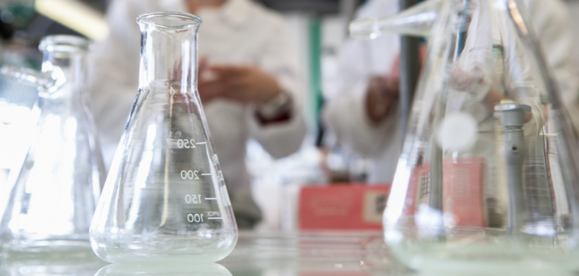 Des fioles Erlenmeyer dans un laboratoire de chimie. Des gens en blouse en arrière-plan dont on ne voit que les mains.