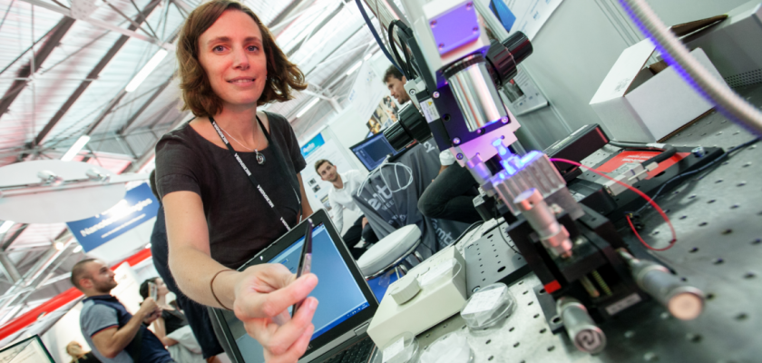 Nadège Courjal sur le stand de Micronora. Elle tient une pince avec un tout petit objet, à côté d'elle un microscope et en arrière plan des gens en train de discuter.