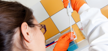 Une jeune femme de dos en blouse et gants oranges manipule une pipette.