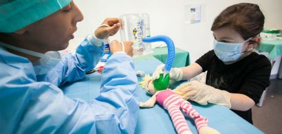 Une petite fille porte un masque et des gants en latex. Elle applique un masque et un tuyau sur la bouche de sa poupée pendant qu'un étudiant en médecine lui explique quelque chose.