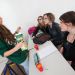 Une jeune fille debout tient un pack de jus de fruit et parle à d'autres jeunes filles assises dans une salle de cours.  .