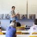 Un homme parle en langue des signes devant un tableau dan une salle de classe où sont assises trois femmes.