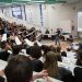 Un amphithéâtre plein de lycéens. Un doctorant fait de grands gestes devant eux. Au fond, 4 personnes du jury.