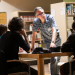 Un homme debout appuyé sur une table où des gens réfléchissent dans une bibliothèque.