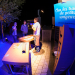 La nuit, un stand avec un jeune homme en train d'expliquer quelque chose à deux autres. La lumière est bleutée. Devant le stand un panneau indique "sur les traces des petits rongeurs"